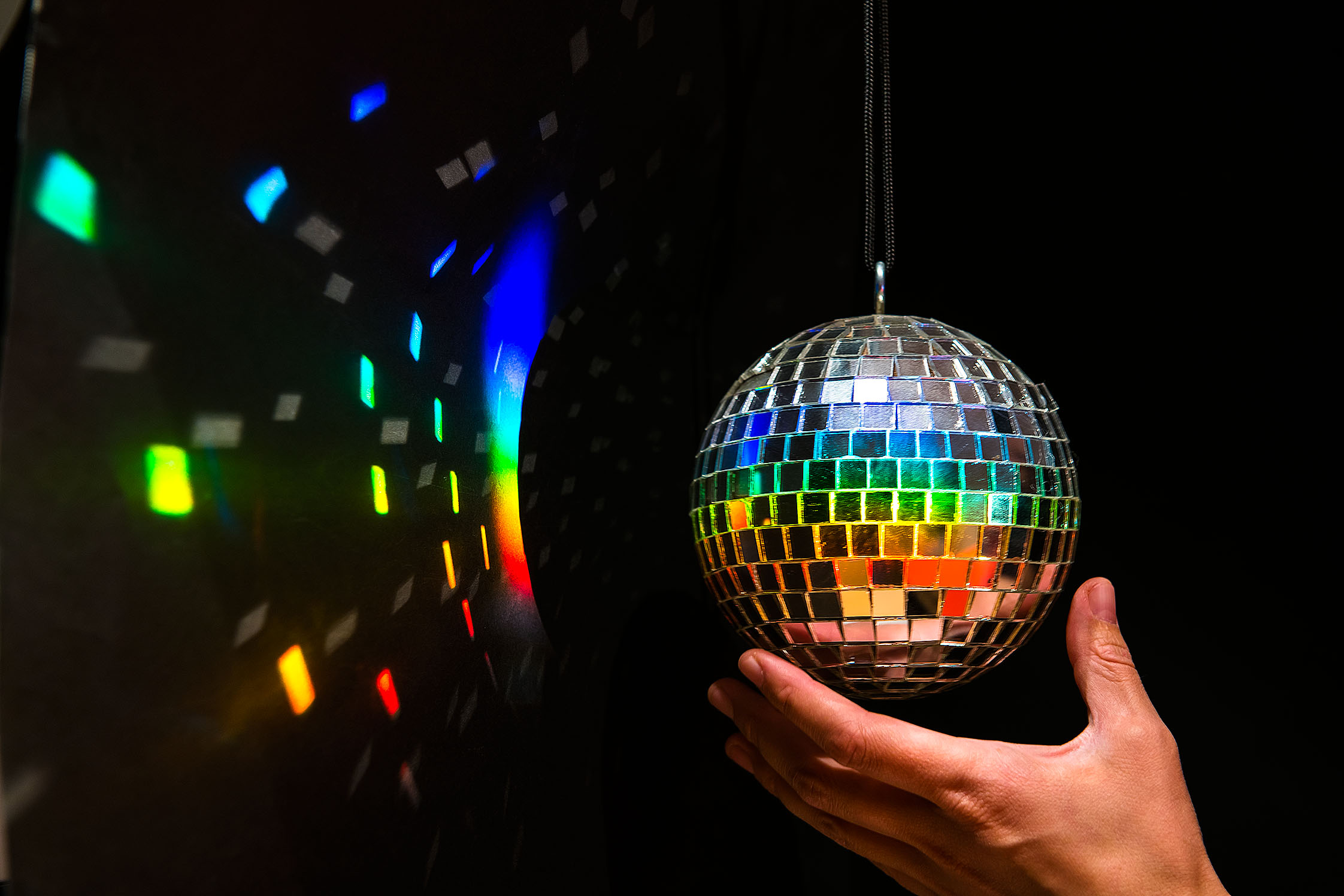 Eine kleine Diskokugel hängt vor dunklem Hintergrund. Auf ihr leuchten die Spektralfarben, die farbigen und weißen Reflektionen von den einzelnen Spiegeln der Diskokugel leuchten auf dem Hintergrund. Eine Hand greift von unten an die Kugel.