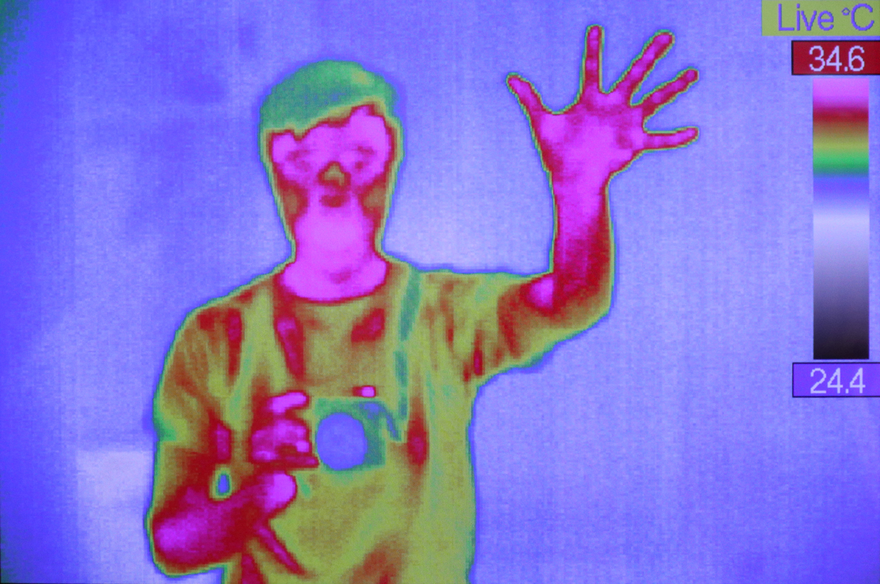 Ein Bildschirm zeigt den Oberkörper einer Person. Sie ist in verschiedenen Farben dargestellt, die der Wärmeverteilung entsprechen. Rechts im Bild befindet sich eine Farbskala, die anzeigt, welche Farbe welcher Temperatur entspricht. Die Person winkt in die Kamera, während sie mit der anderen Hand etwas vor ihren Körper hält.