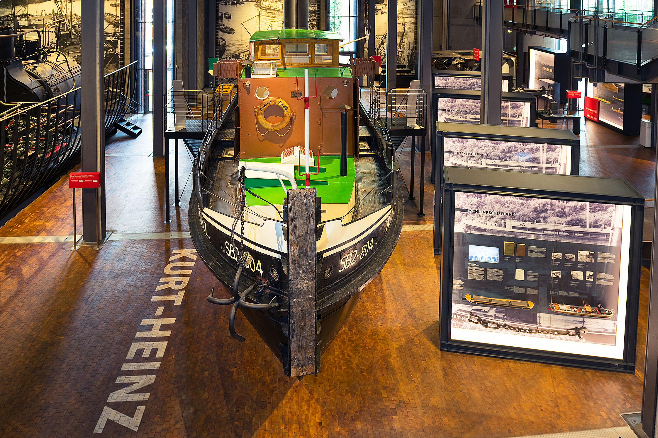 Im Ausstellungsraum steht der Dampfschlepper KURT-HEINZ. Links daneben ist die Bodenbeschriftung angebracht, rechts stehen mehrere große Leuchtvitrinen hintereinander.