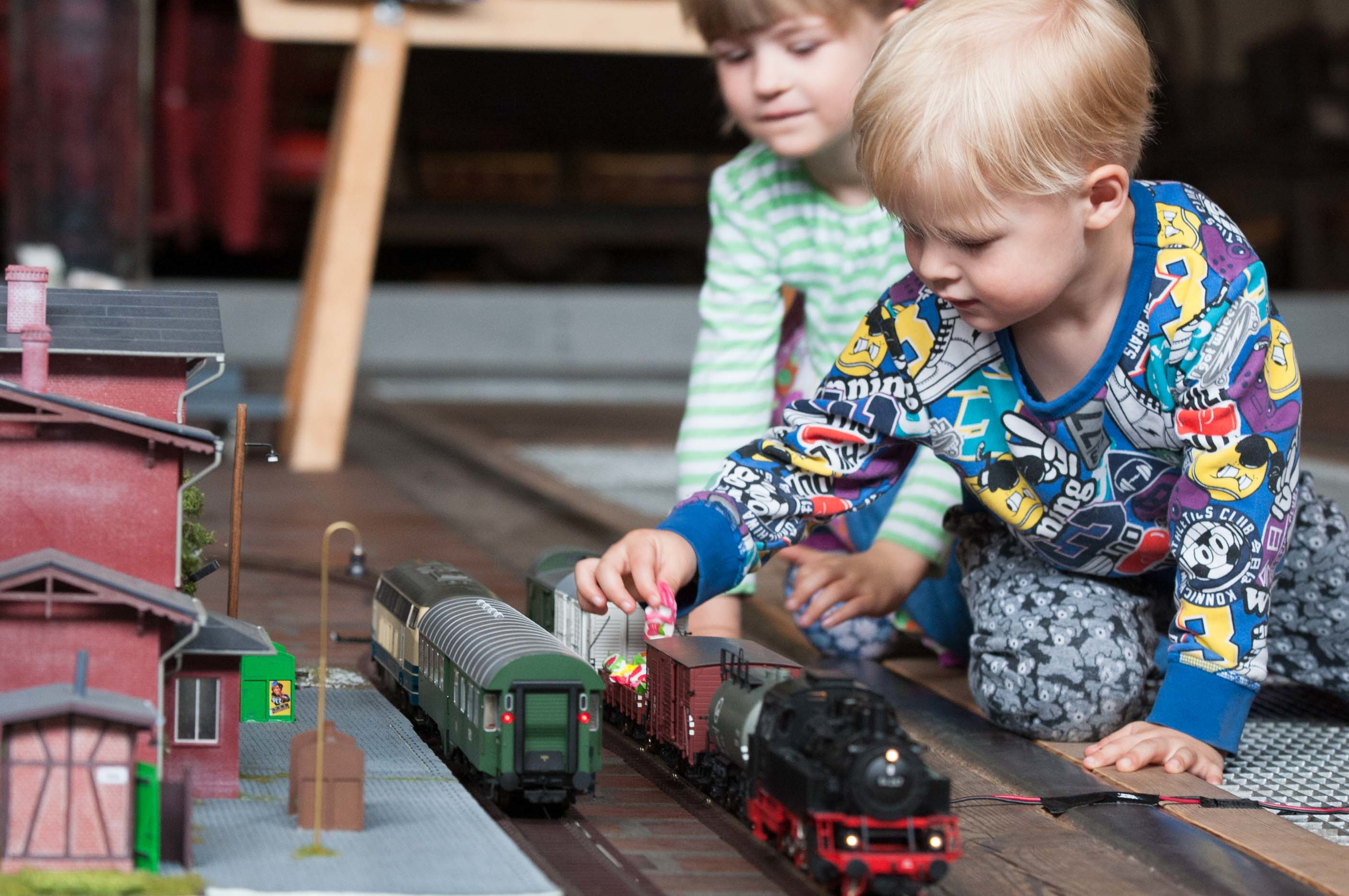 Zwei Kinder sitzen neben einer Spur-0-Modelleisenbahn auf einem Teppich. Auf den Gleisen stehen eine schwarz-rote Lokomotive und mehrere Anhänger. Eines der Kinder belädt einen offenen Anhänger mit kleinen, buntverpackten Süßigkeiten.