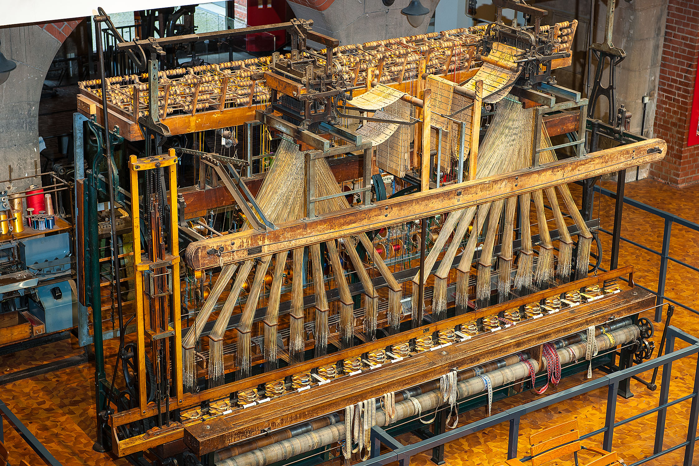 Ein großer, historischer Bandwebstuhl aus Holz und Metall. Auf der Oberseite des Webstuhls sind zwei Jacquardaufsätze mit Lochkarten zu erkennen, mit denen das Muster des späteren Stoffs vorgegeben wird.