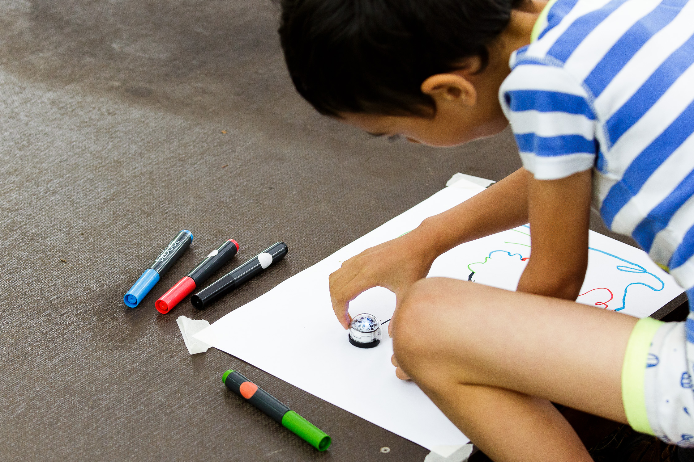 Ein kleiner Junge lässt einen Miniroboter auf einer Linie entlang fahren, die er vorher auf ein Stück Papier gezeichnet hat.