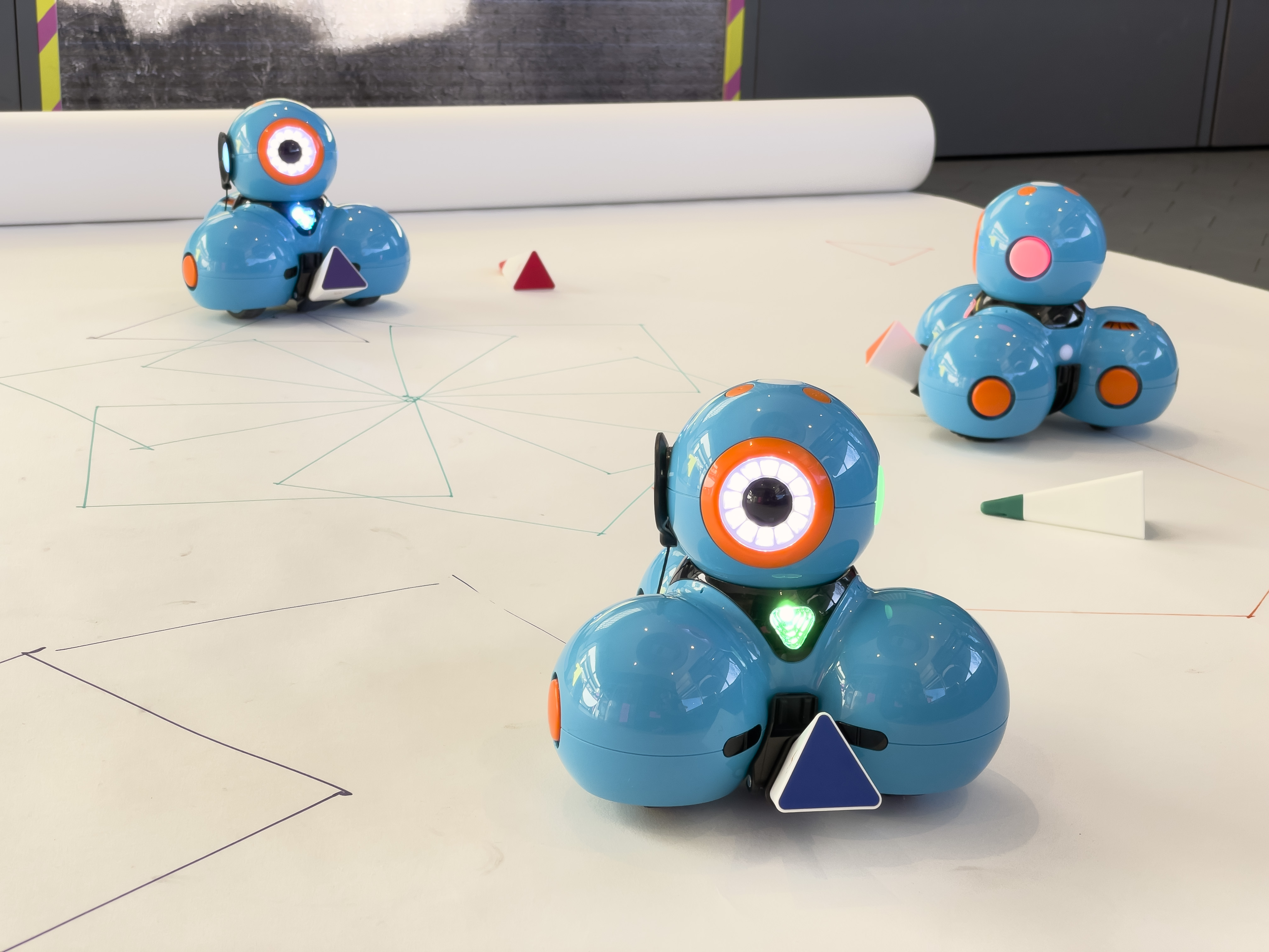 Drei blaue Roboter stehen auf weißem Untergrund. Auf dem Untergrund sind geometrische Formen gezeichnet.