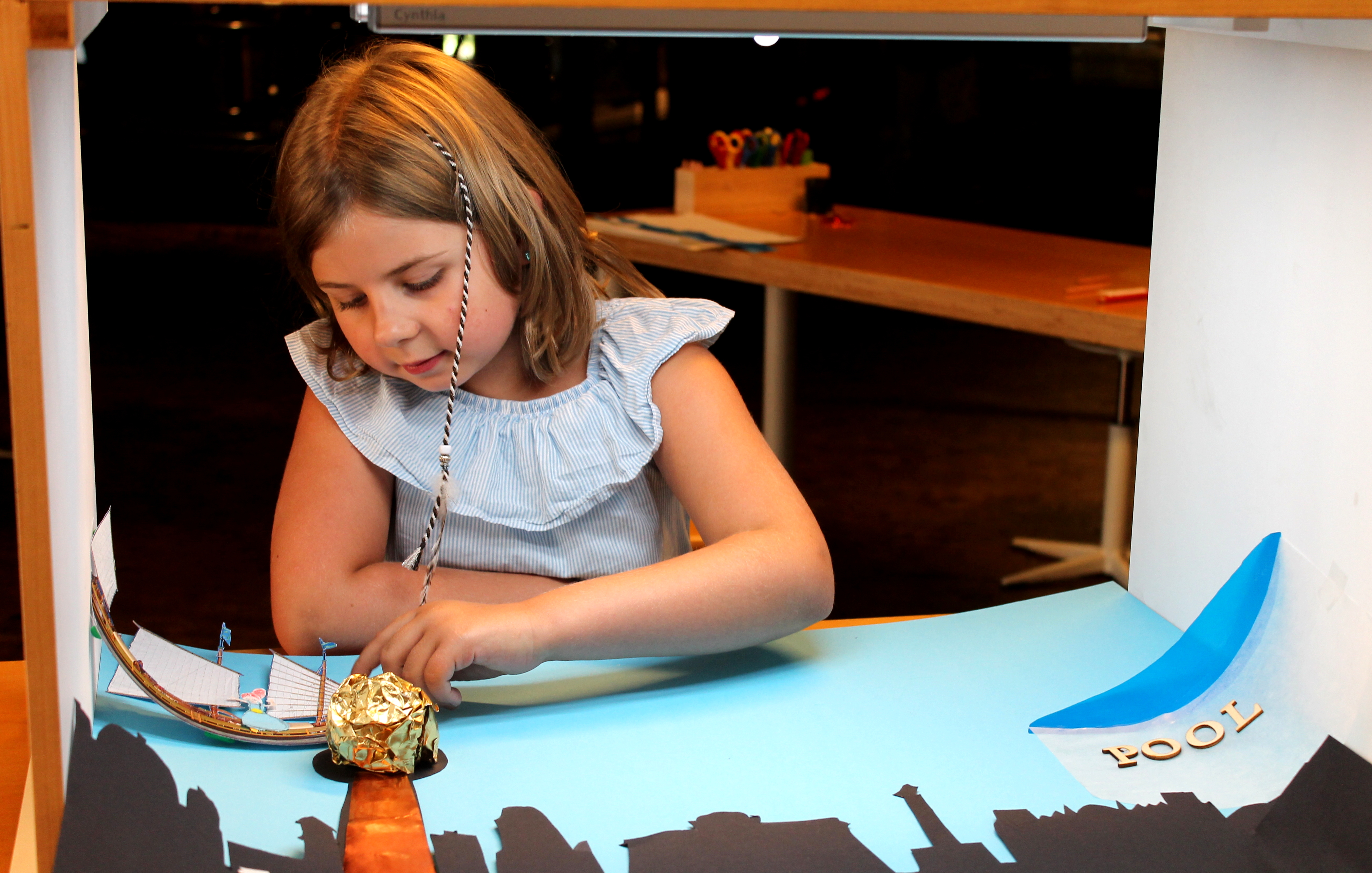 Ein Mädchen sitzt an einem Tisch, auf dem eine Kiste mit zwei offenen Seiten steht. In der Kiste liegen eine ausgeschnittene Stadtsilhouette und ein Schiff aus Papier auf einem blauen Hintergrund. Das Mädchen verschiebt das Schiff mit der linken Hand.