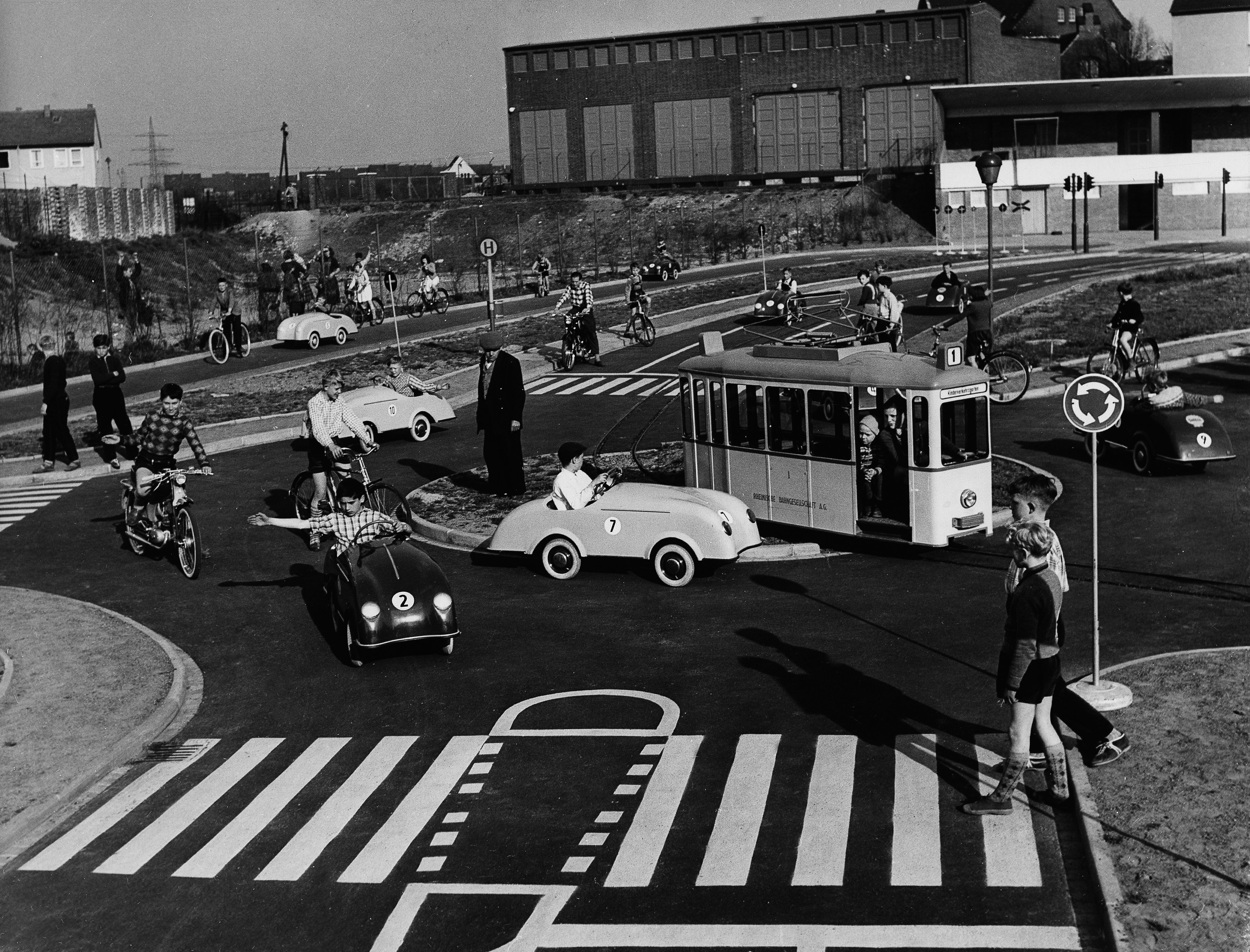 Schwarz-Weiß-Aufnahme, Kinder auf einem Verkehrsparcours in unterschiedlichen Fahrzeugen, im Vordergrund überqueren zwei Kinder einen Zebrastreifen.
