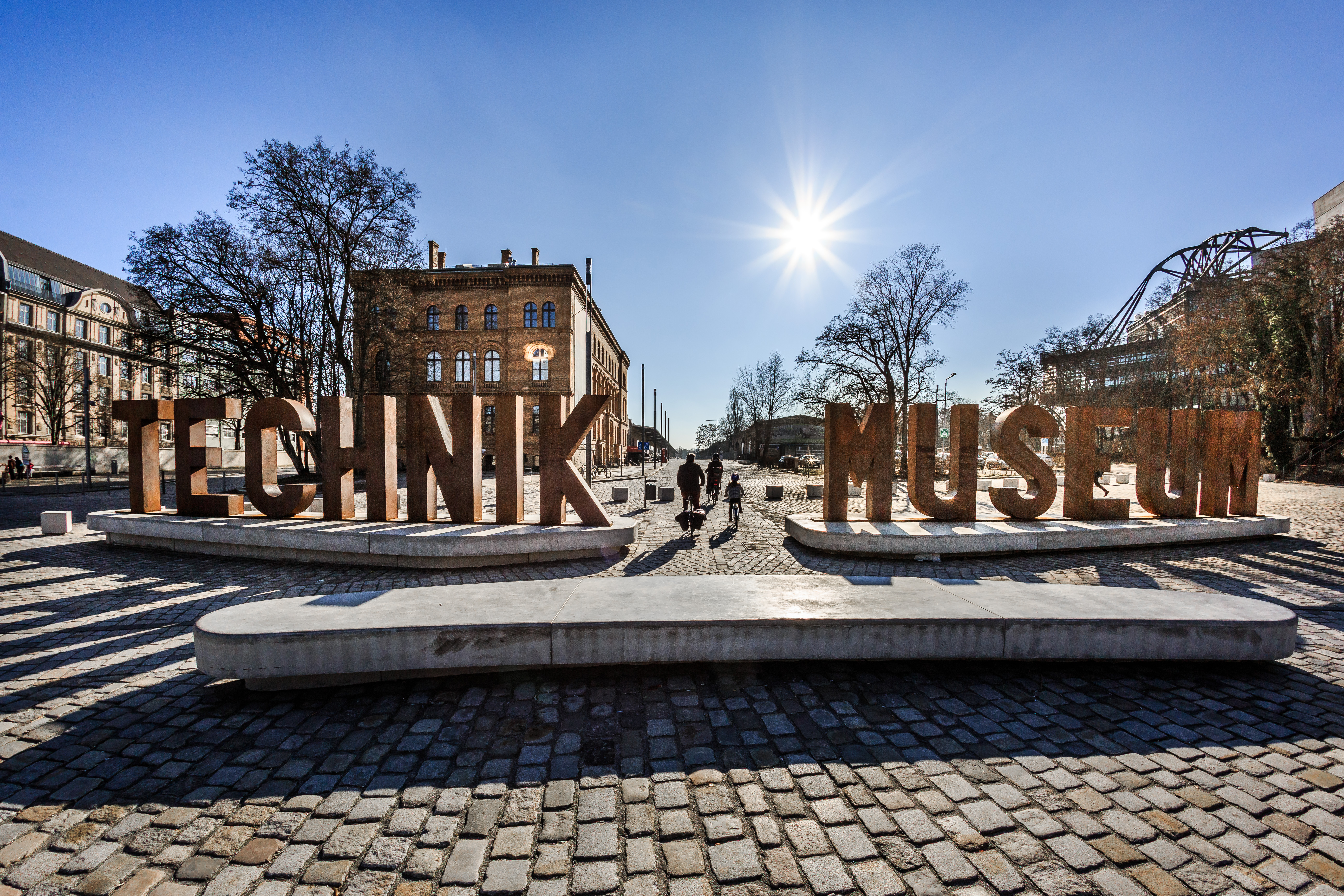 Auf einem gepflasterten Platz sieht man eine aus Metallbuchstaben erstellte Wortskulptur, die sich „Technik Museum“ liest. Im Hintergrund ein Backsteingebäude.