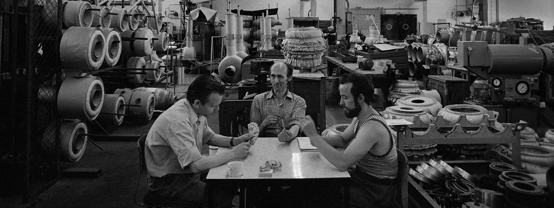 Auf dem Foto sieht man drei Männer, die an einem Tisch mitten in einer riesigen Werkshalle sitzen und Karten spielen.