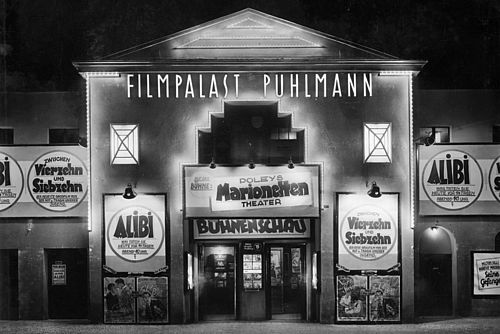Schwarzweiß-Foto: Beleuchtetes Gebäude mit großem Schriftzug „Filmpalast Puhlmann“. An der Fassade sind viele Plakate und Banner angebracht, die für verschiedene Filme werben.