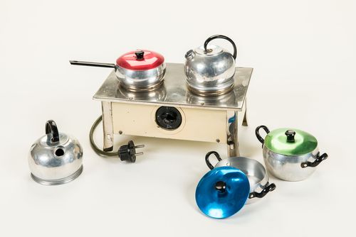 This brightly enameled miniature stove is operated electrically. The stove is equipped with a power cord and plug and has a rotary switch at the front for turning it on and off. Cooking was carried out on two hotplates with the accompanying metal cookware: two kettles, a saucepan with a red lid and two cooking pots with a blue and a green lid, respectively.