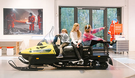 Three children sit on a black and yellow snowmobile in an exhibition hall..