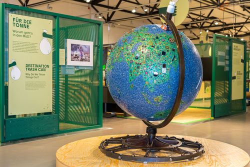 This image shows a large globe in the exhibition foyer. Its surface represents the world in a mosaic of electronic waste: the seas are made of small blue circuit board parts, the forests from green circuit boards, and the mountains from brown cables. 