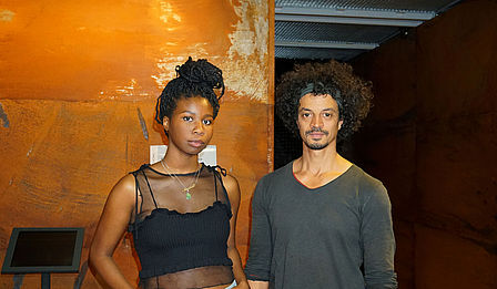 Die nigerianisch-amerikanische Künstlerin Monilola Ilupeju und der deutsch-nigerianische Künstler Philip Kojo Metz stehen vor der rostbraunen Wand des Moduls zum Versklavungshandel im Deutschen Technikmuseum.