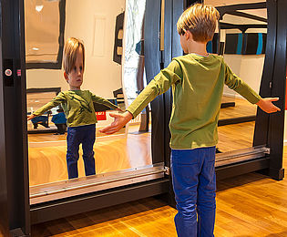 Ein Kind steht vor einem gekrümmten Spiegel und betrachtet sein verzerrtes Spiegelbild.