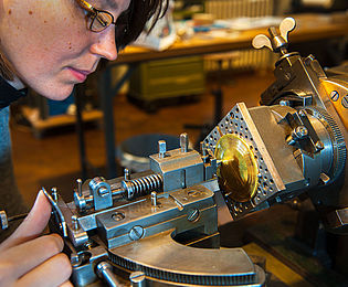 Eine Frau bedient eine Guillochier-Maschine. Mit der rechten Hand bewegt sie die Graviernadel der Maschine über das eingespannte Metallstück. 