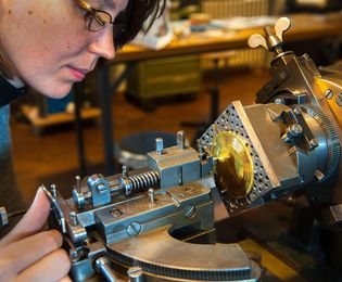 A woman operates a Guilloché machine. Her right hand moves the machine’s engraving needle over a piece of metal.