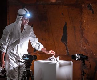A dark-skinned woman who is dressed in a white protective suit with a headlamp reaches out with one hand to touch a pile of dust lying on a pedestal. A brown metal wall can be seen in the background.