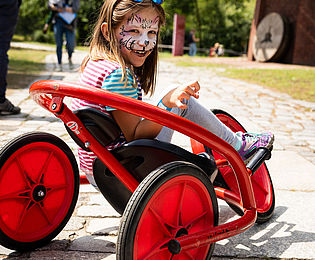 A little girl is sitting on a red vehicle. It is made up as a cat.