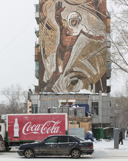 Ein Auto und ein Lastwagen mit Coca-Cola-Werbung fahren über eine verschneite Straße. Im Hintergrund steht ein Hochhaus mit Wandmosaik, auf dem ein überlebensgroßer Kosmonaut in den Himmel schwebt.