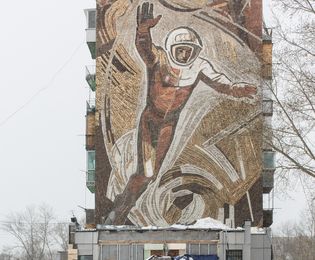 A car and a truck bearing a Coca-Cola advertisement are driving along a snow-covered road. In the background is a high-rise building with a mosaic mural depicting a larger-than-life cosmonaut floating up to the sky.