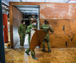 Several people in green overalls are removing materials from a brown metal cubical structure.