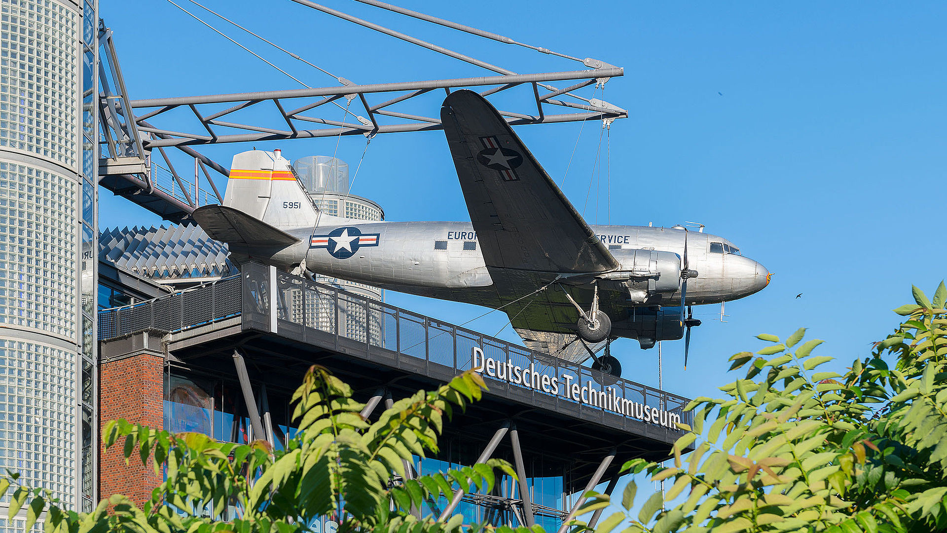 Ein Neubaugebäude mit Glasfassade. An einer Stahlkonstruktion auf dem Dach hängt ein Flugzeug. Unter dem Flugzeug hängt eine weiße Leuchtschrift „Deutsches Technikmuseum“.