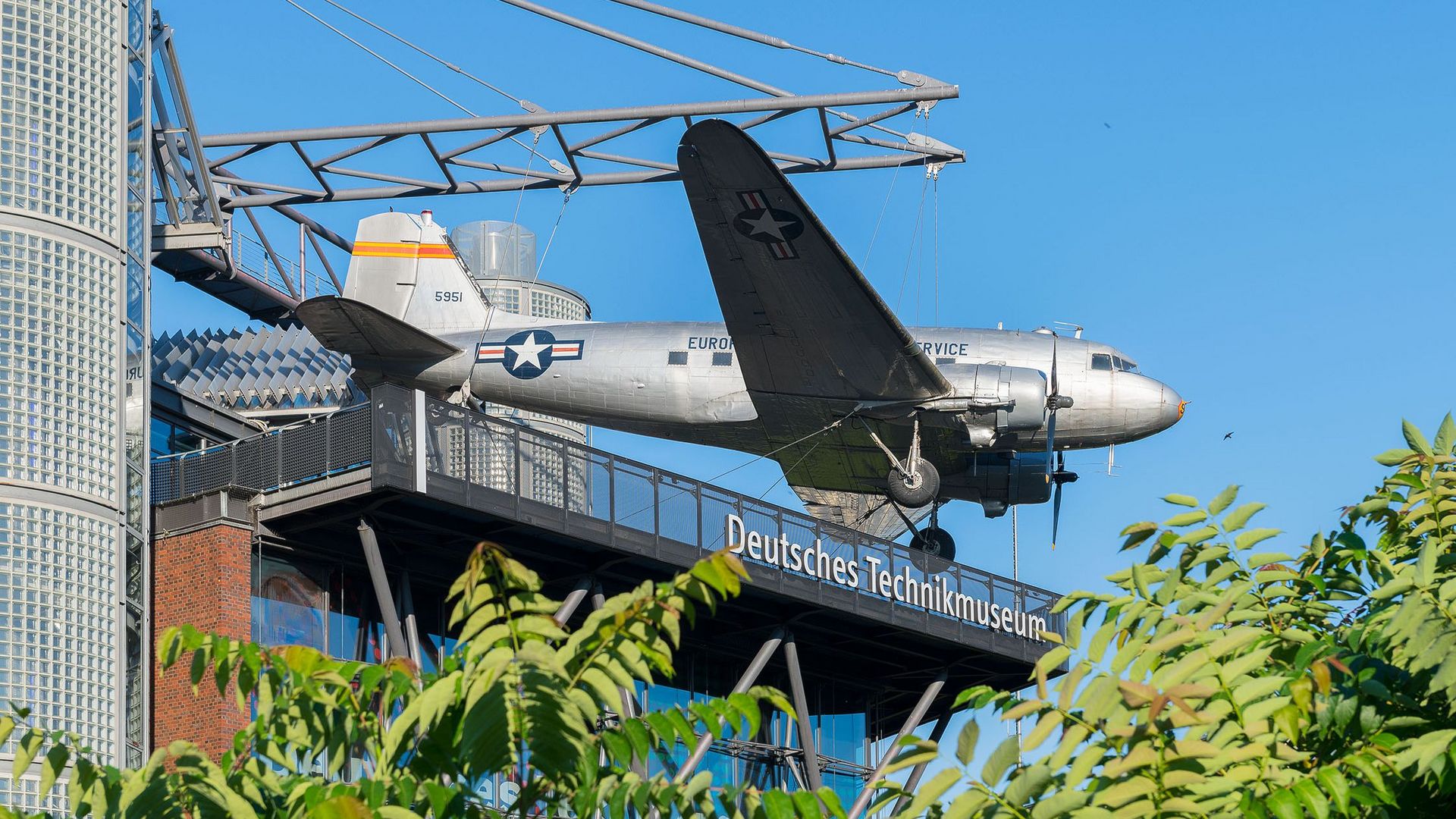Ein Neubaugebäude mit Glasfassade. An einer Stahlkonstruktion auf dem Dach hängt ein Flugzeug. Unter dem Flugzeug hängt eine weiße Leuchtschrift „Deutsches Technikmuseum“.