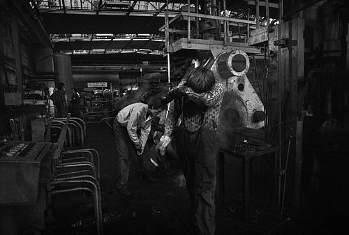 Das Foto zeigt zwei Arbeiter, die mit schweren Arbeitsgeräten in einer Werkhalle hantieren und offensichtlich stark erschöpft sind.z