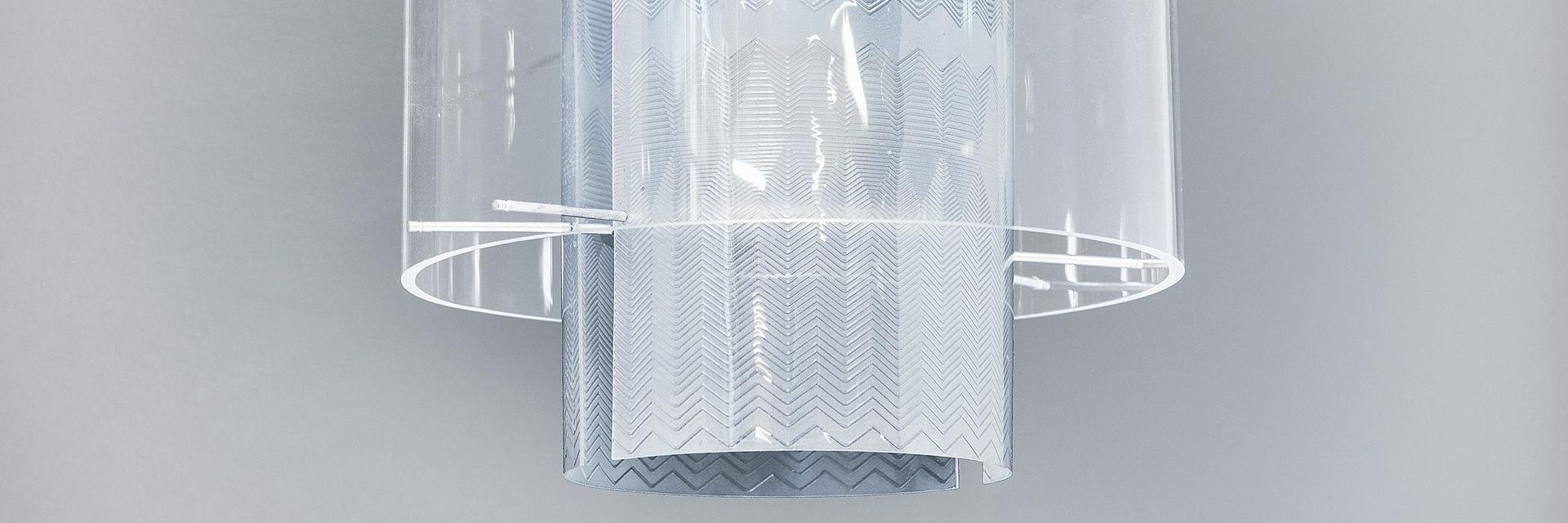 The lamp is hung on a white cord. The light bulb is encased with transparent, rounded plexiglass panes that are engraved with decorative zigzag lines. 