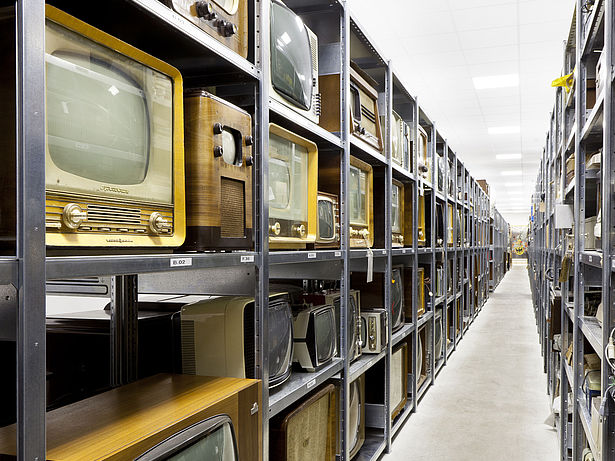 Ein schmaler Gang zwischen langen Metallregalen. In den Regalen stehen viele historische Fernsehgeräte mit Holzgehäuse.