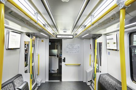 Blick in den Innenraum eines U-Bahnwagens. Der Eindruck ist hell und groß. Die Sitze sind blau-schwarz gemustert, die Haltestangen sind gelb.