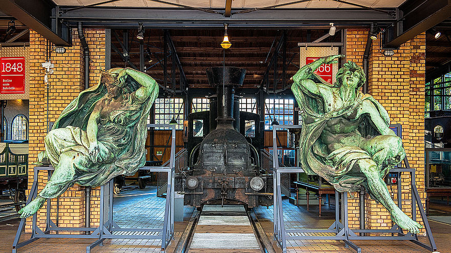 Zwei große, grün angelaufene Bronze Figuren stehen links und rechts vor den gelben Backsteinsäulen der historischen Lokschuppen. Zwischen Ihnen steht eine schwarze Lokomotive.