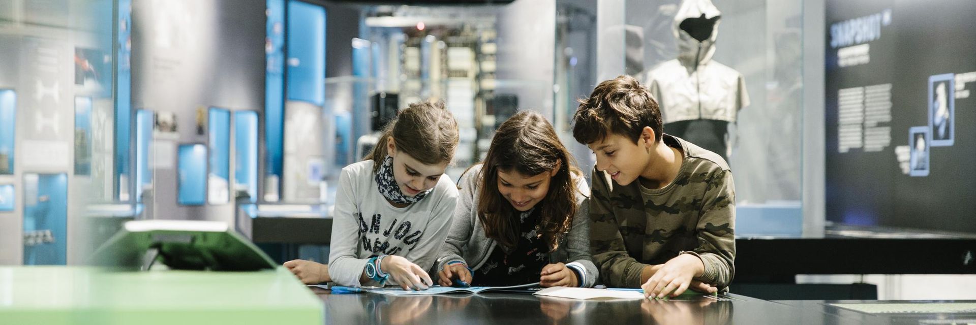 Drei Kinder stehen, konzentriert über ein Heft gebeugt, in der Ausstellung. Im Hintergrund sieht man Vitrinen, Objekte und Wandtexte.