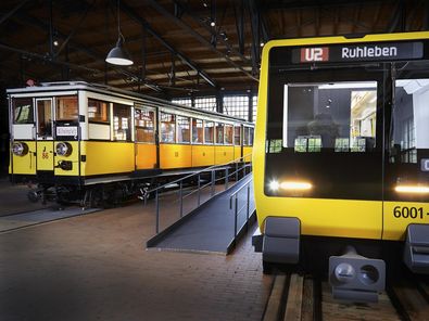Blick auf zwei nebeneinander stehende U-Bahnwagen. Beide sind gelb. Der Wagen rechts sieht sehr modern aus, der Wagen links ist ein historisches Fahrzeug.