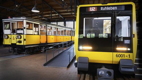 Blick auf zwei nebeneinander stehende U-Bahnwagen. Beide sind gelb. Der Wagen rechts sieht sehr modern aus, der Wagen links ist ein historisches Fahrzeug.