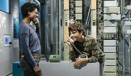 A woman and a boy stand at a table in the exhibition with two corded telephones on it. Both of them are talking on a phone, holding a receiver up to their face.
