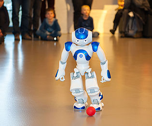 Der weiß-blaue Roboter NAO spielt mit einem kleinen roten Ball. Der etwa 60 Zentimeter große humanoide Roboter geht auf den Ball zu und versucht ihn zu treten. Im Hintergrund verfolgen Kinder und Erwachsene die Vorführung.