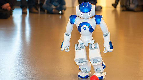 Der weiß-blaue Roboter NAO spielt mit einem kleinen roten Ball. Der etwa 60 Zentimeter große humanoide Roboter geht auf den Ball zu und versucht ihn zu treten. Im Hintergrund verfolgen Kinder und Erwachsene die Vorführung.