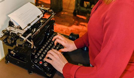 A young woman stands at a black typewriter. A piece of white paper is inserted in the typewriter. The woman’s fingers are at the keyboard. She is typing.