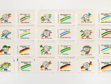 24 white, square sugar packets are lined up chronologically in the order of their World Cup titles. They are imprinted with such motifs as soccer balls wearing traditional national hats or waving national flags as well the national colors of the respective World Cup Champions and advertising slogans such as "Sugar, a thing of perfection" or "Sugar boosts endurance."