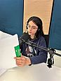 Eine junge Person mit braunen schulterlangen Haaren und einer Brille sitzt in einem Tonstudio vor einem Mikrofon und hält ein Skript in der Hand.