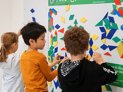 Drei Kinder stehen vor einer weißen Magnetwand und legen symmetrische Figuren aus verschiedenen bunten Formen.
