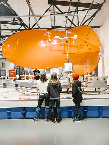 Ein großer orangefarbener Fesselballon hängt in einer Ausstellungshalle. Drei Kinder stehen davor und betrachten ihn.