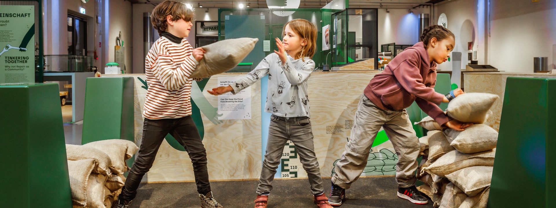Drei Kinder reparieren in einer Ausstellungshalle einen Spielzeug-Deich aus Sandsäcken. Sie bilden eine Kette und reichen sich die Säcke, um eine Lücke im Deich zu flicken.