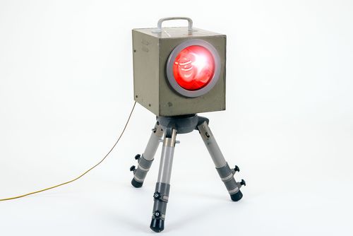 A rectangular, elongated metal flash unit is mounted on a tripod. The legs are painted grey while the housing is somewhat darker and melds into an olive green color. There is a red round glass disk on the front of the flash unit.