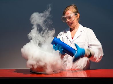 Hinter einem Pult mit roter Oberfläche steht eine Frau in einem weißen Laborkittel. Sie trägt eine Schutzbrille und blaue, dicke Handschuhe. Sie hält eine blaue, Isolierkanne leicht gekippt in den Händen. Unter der Öffnung befindet sich eine Wolke aus weißem Nebel, der bis auf die Tischplatte reicht.