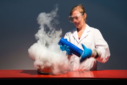 Hinter einem Pult mit roter Oberfläche steht eine Frau in einem weißen Laborkittel. Sie trägt eine Schutzbrille und blaue, dicke Handschuhe. Sie hält eine blaue, Isolierkanne leicht gekippt in den Händen. Unter der Öffnung befindet sich eine Wolke aus weißem Nebel, der bis auf die Tischplatte reicht.