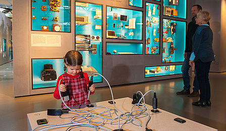 Ein kleines Mädchen spielt mit einem Steckspiel aus übergroßen Computer- und Telefonverbindungssteckern. Hinter ihr stehen zwei Museumsbesucher und schauen auf die Gegenstände in einer hell erleuchteten Vitrinenwand.