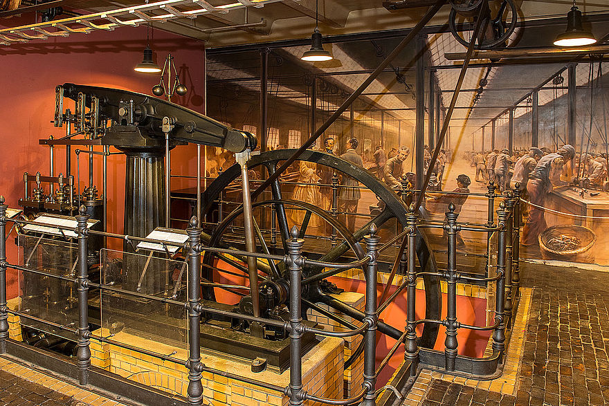 The central transmission shaft is enclosed by a railing. The smaller drive wheel with the transmission belt and the larger flywheel can be seen behind it. The pistons of the steam engine are in the foreground. In the background, there is a mural depicting a historical machine shop.