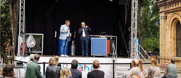 Two men stand on an open-air stage and speak into microphones. People are sitting in the foreground and listening to them. In the foreground people are also sitting and listening. 