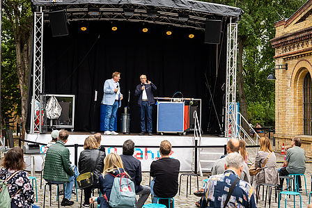 Zwei Männer stehen auf einer Open-Air Bühne und sprechen in Mikrofone. Im Vordergrund sitzen Menschen und hören ihnen zu.
